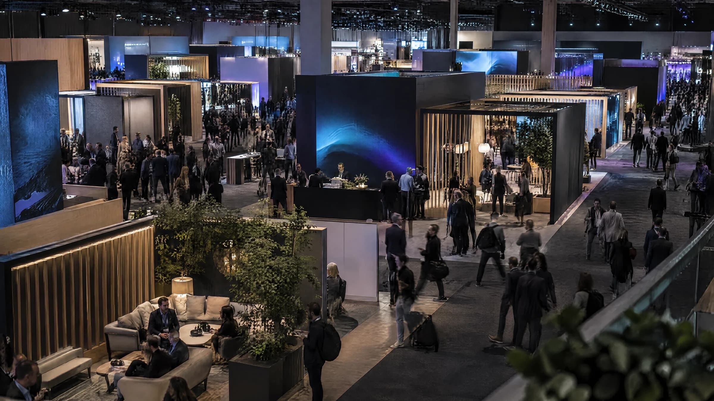 An expo hall from above: dark booths with lit screens, a lounge area and visitors walking the aisles.