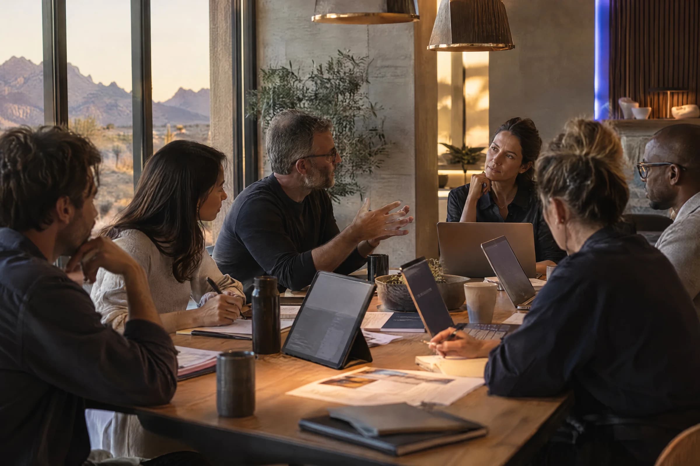 A small group around a wooden table with laptops and tablets, desert mountains visible through the window.